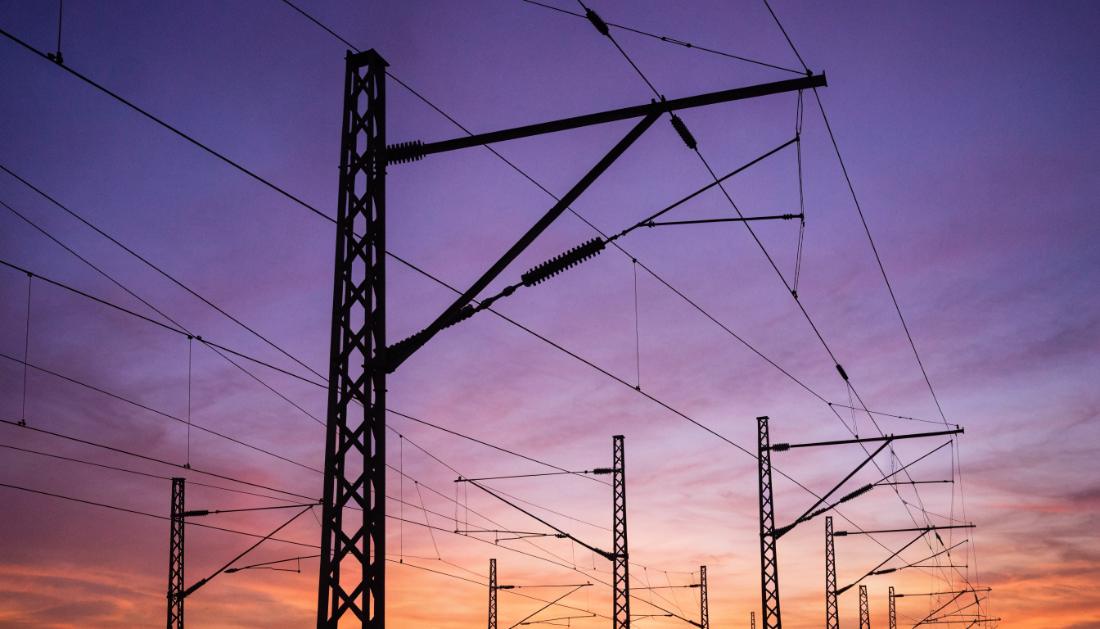 Oberleitungsmasten und Fahrdraehte als Silhouette vor orange-violettem Abendhimmel