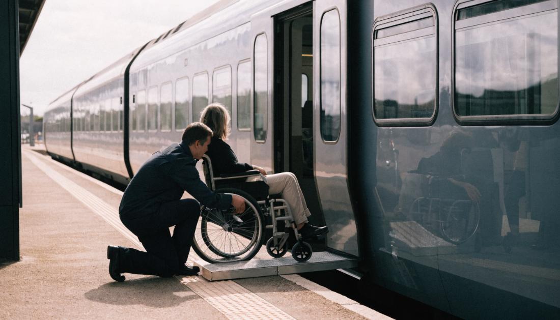 Ein Mitarbeiter legt eine mobile Rampe zwischen Bahnsteigkante und Zugt&uuml;r, eine Rollstuhlfahrerin wartet daneben