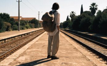Ein junger Reisender mit grossem Rucksack blickt auf einem sonnigen Bahnsteig die Gleise entlang