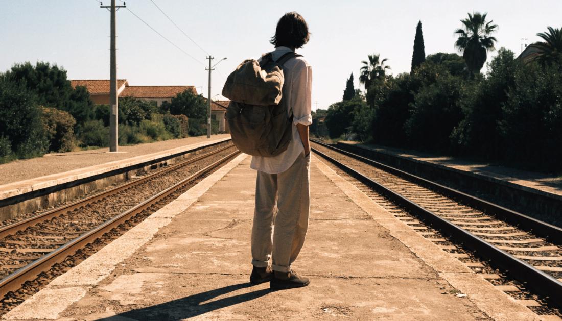Ein junger Reisender mit gro&szlig;em Rucksack blickt auf einem sonnigen Bahnsteig die Gleise entlang