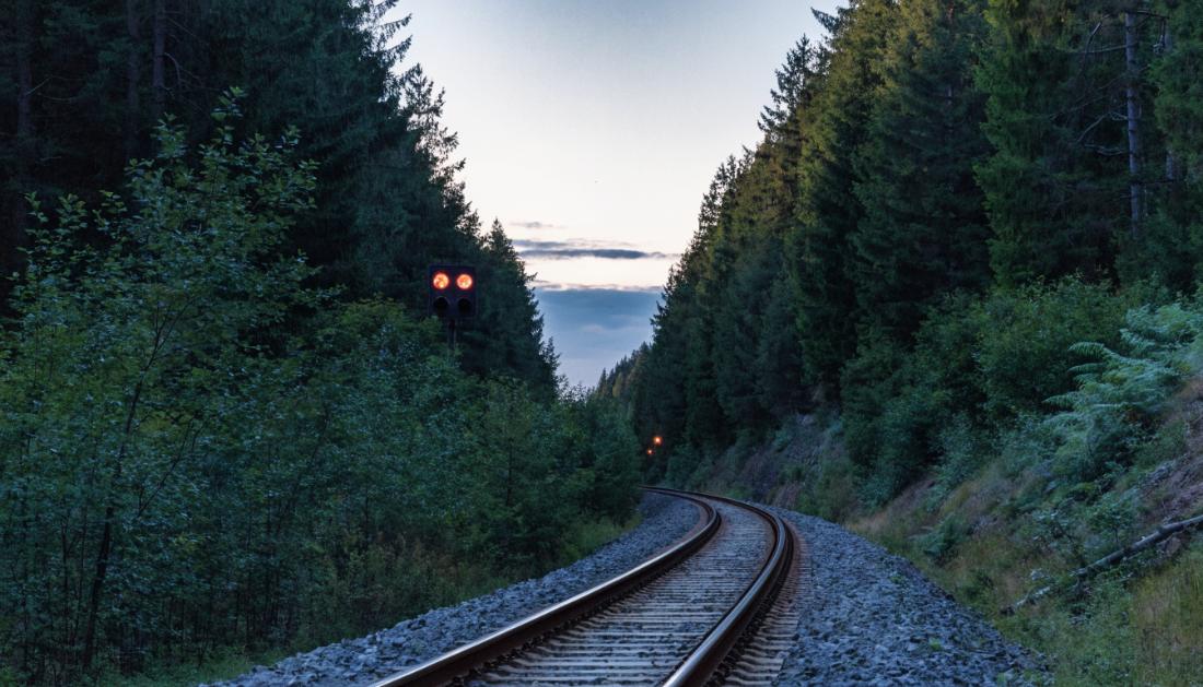 Ein leeres Gleis schlängelt sich in der Dämmerung durch einen ruhigen Wald, Warnlichter leuchten in der Ferne