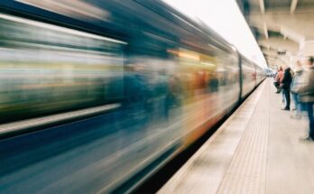 Ein schneller Zug rast mit Bewegungsunschärfe durch einen Bahnsteig, wartende Reisende stehen sicher hinter der Bahnsteigkante
