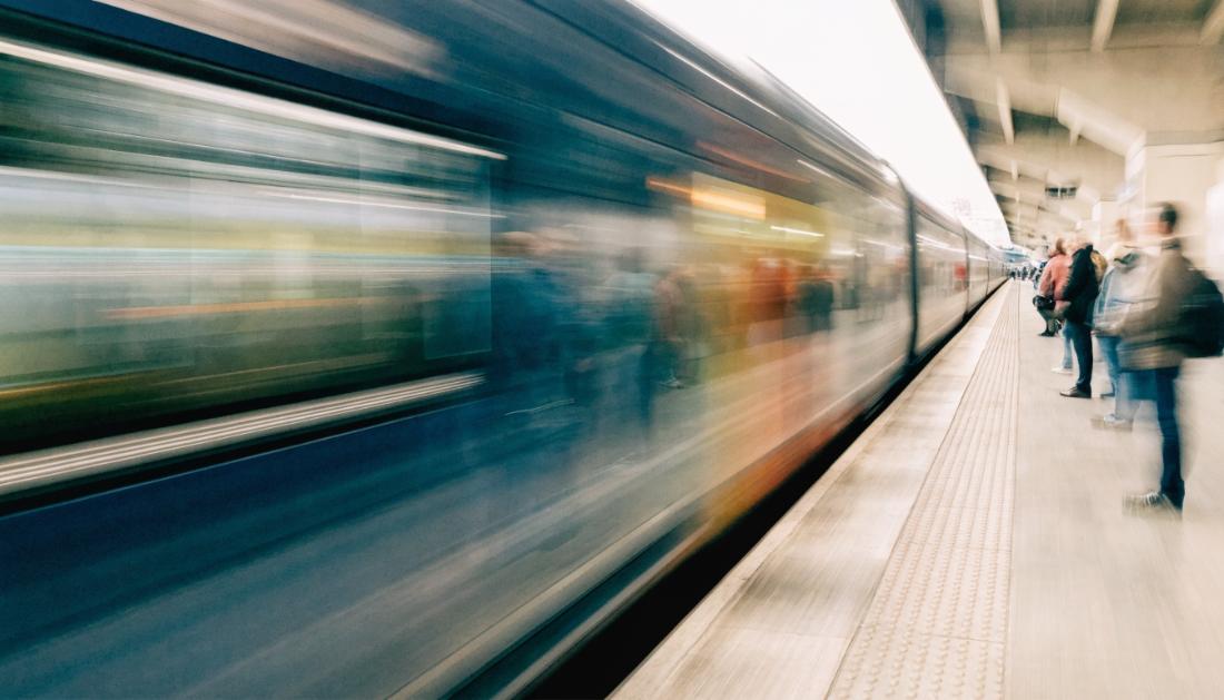Ein schneller Zug rast mit Bewegungsunschärfe durch einen Bahnsteig, wartende Reisende stehen sicher hinter der Bahnsteigkante