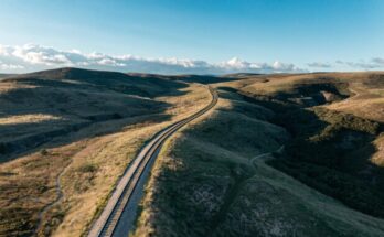 Eine Bahnstrecke schwingt sich elegant durch eine hügelige Landschaft, aus der Vogelperspektive fotografiert