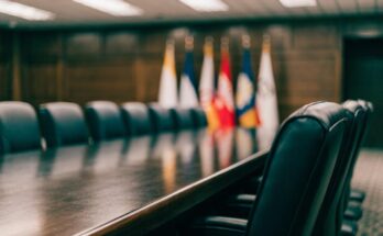 Ein großer formeller Konferenztisch in einem Regierungsraum mit leeren Stühlen und Flaggen im Hintergrund