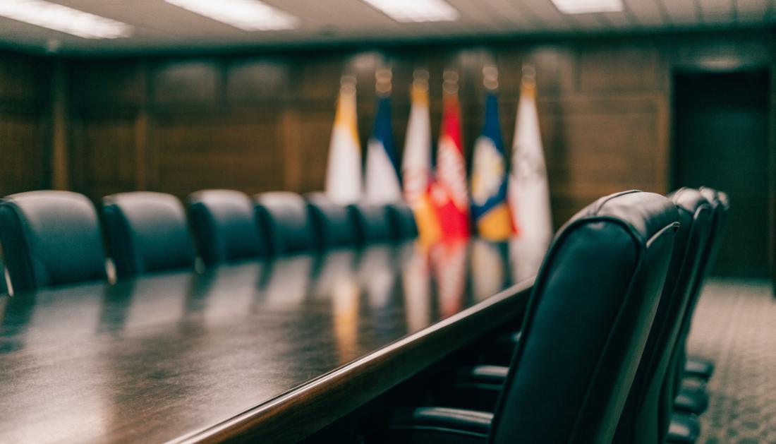 Ein großer formeller Konferenztisch in einem Regierungsraum mit leeren Stühlen und Flaggen im Hintergrund