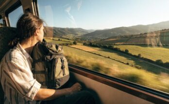 Eine junge Person mit Rucksack sitzt am Zugfenster und blickt auf eine weite europäische Landschaft