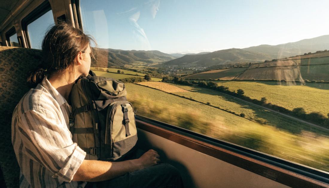 Eine junge Person mit Rucksack sitzt am Zugfenster und blickt auf eine weite europäische Landschaft