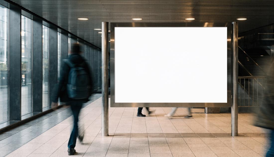 Eine leere helle Plakatfläche in einer Bahnhofshalle, Menschen gehen unscharf vorbei