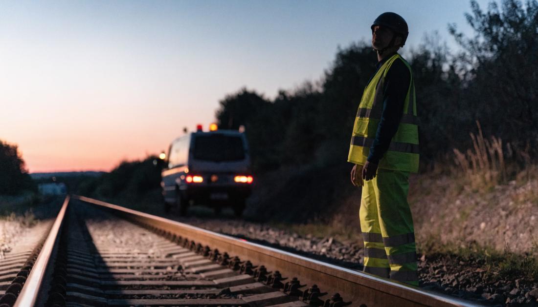 Eine Person in Warnschutzkleidung steht in der Dämmerung neben Bahngleisen
