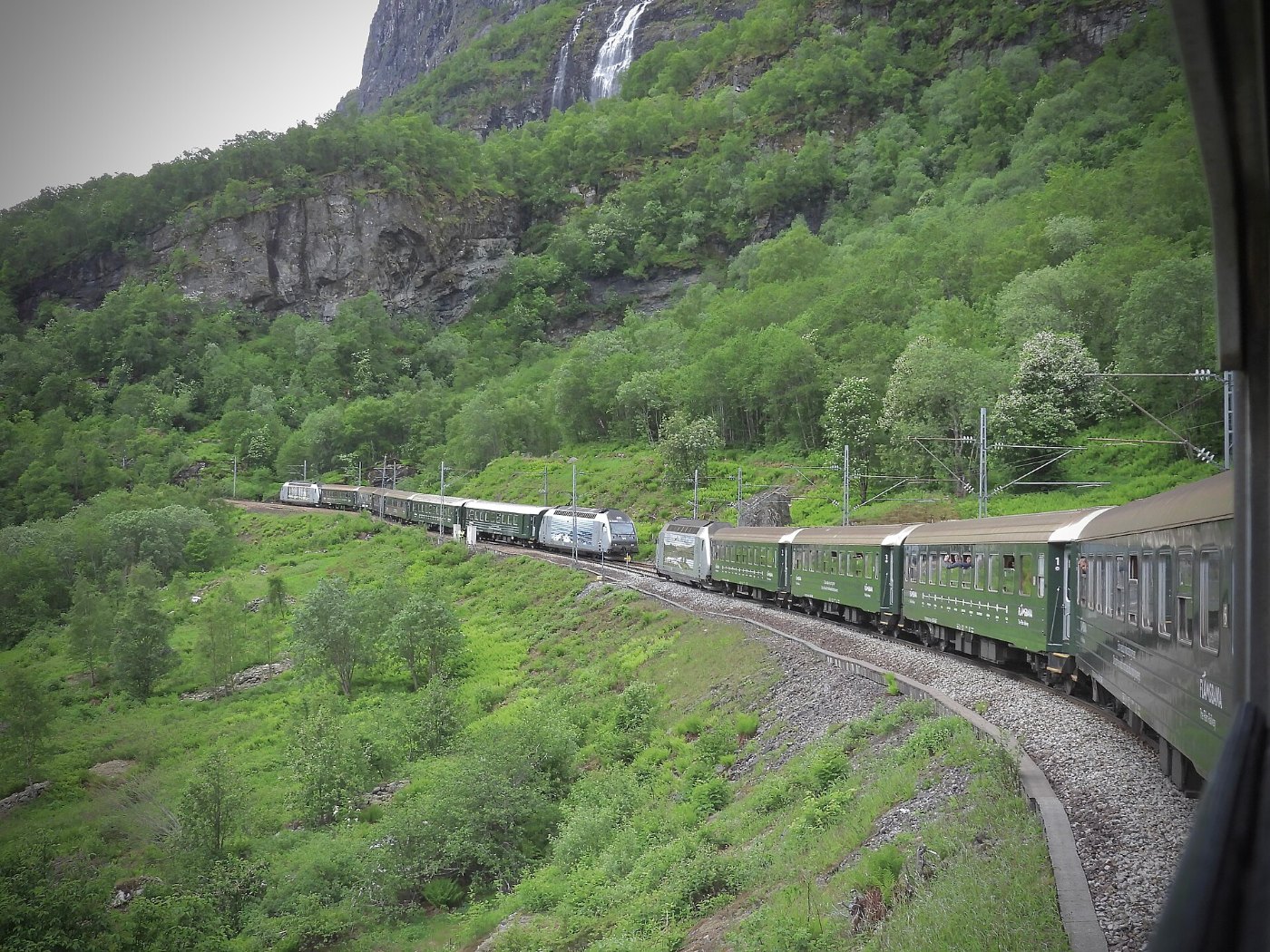 Two Flåmsbana Trains meet and pass as the Brekkefossen Waterfall cascade